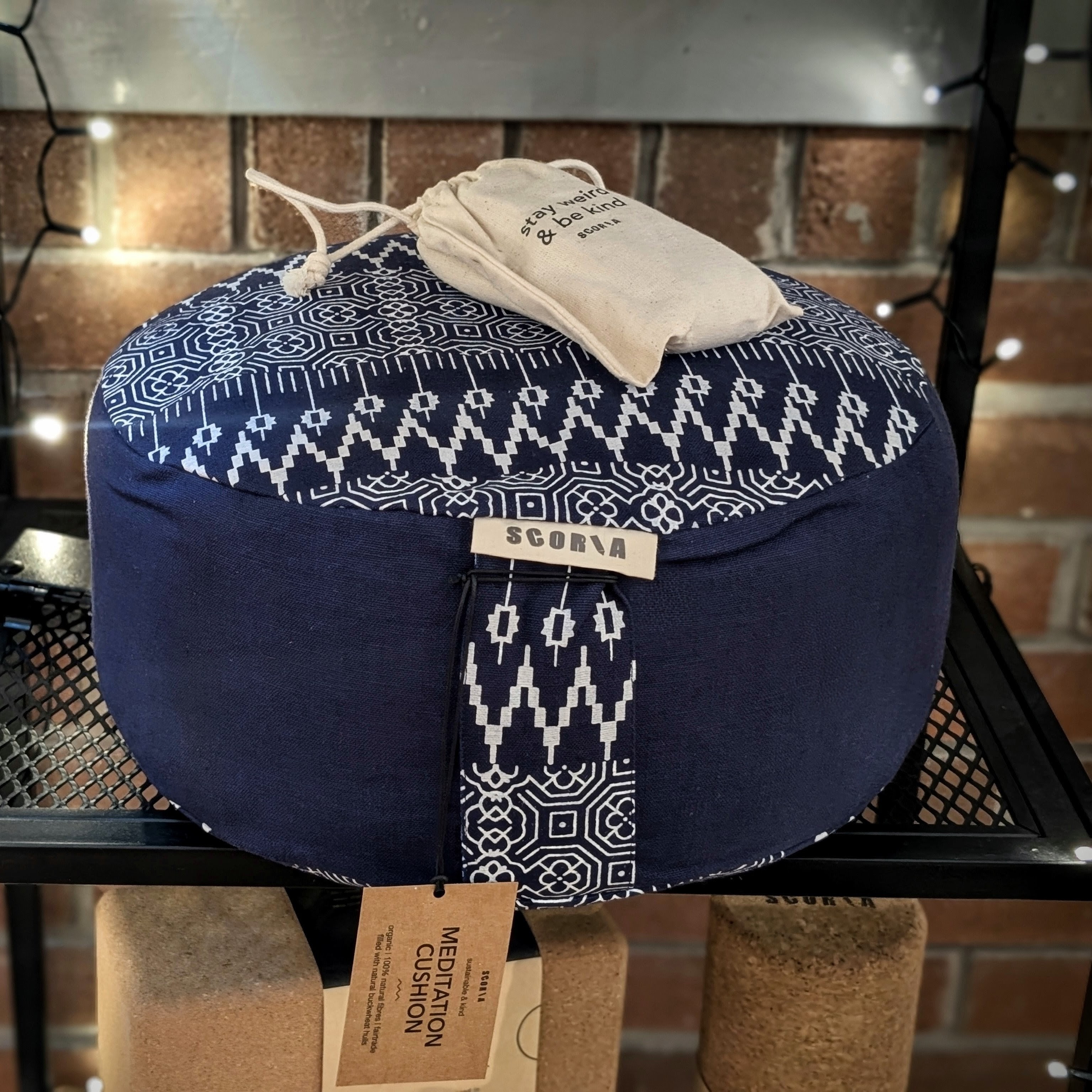 A round meditation cushion in navy blue with a white geometric pattern on top, labeled 'SCORIA'. It is accompanied by a small drawstring bag and a tag that says 'MEDITATION CUSHION'.