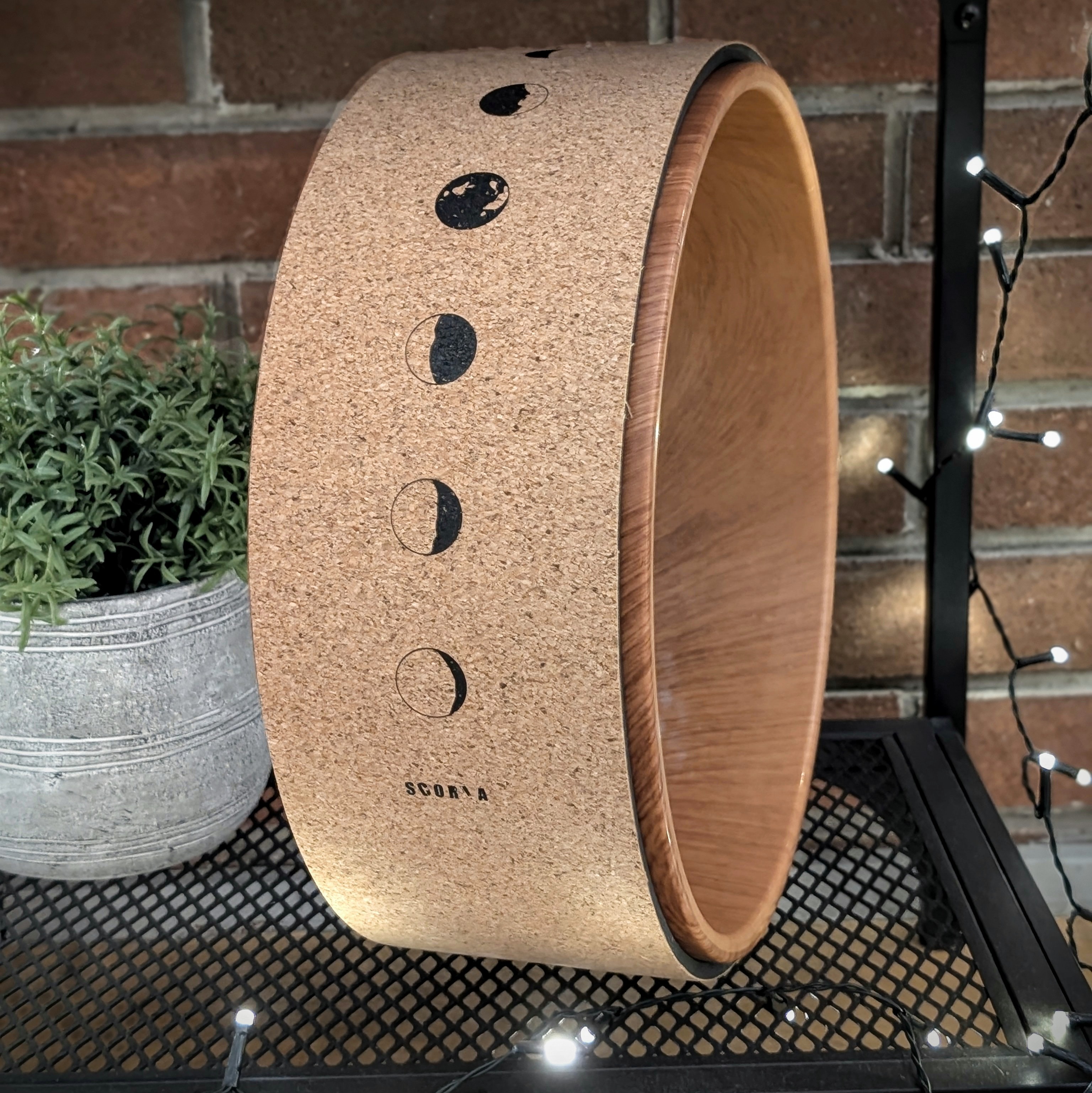 A cork decorative object featuring the phases of the moon illustrated in black, positioned next to a small potted plant and fairy lights on a metal stand.
