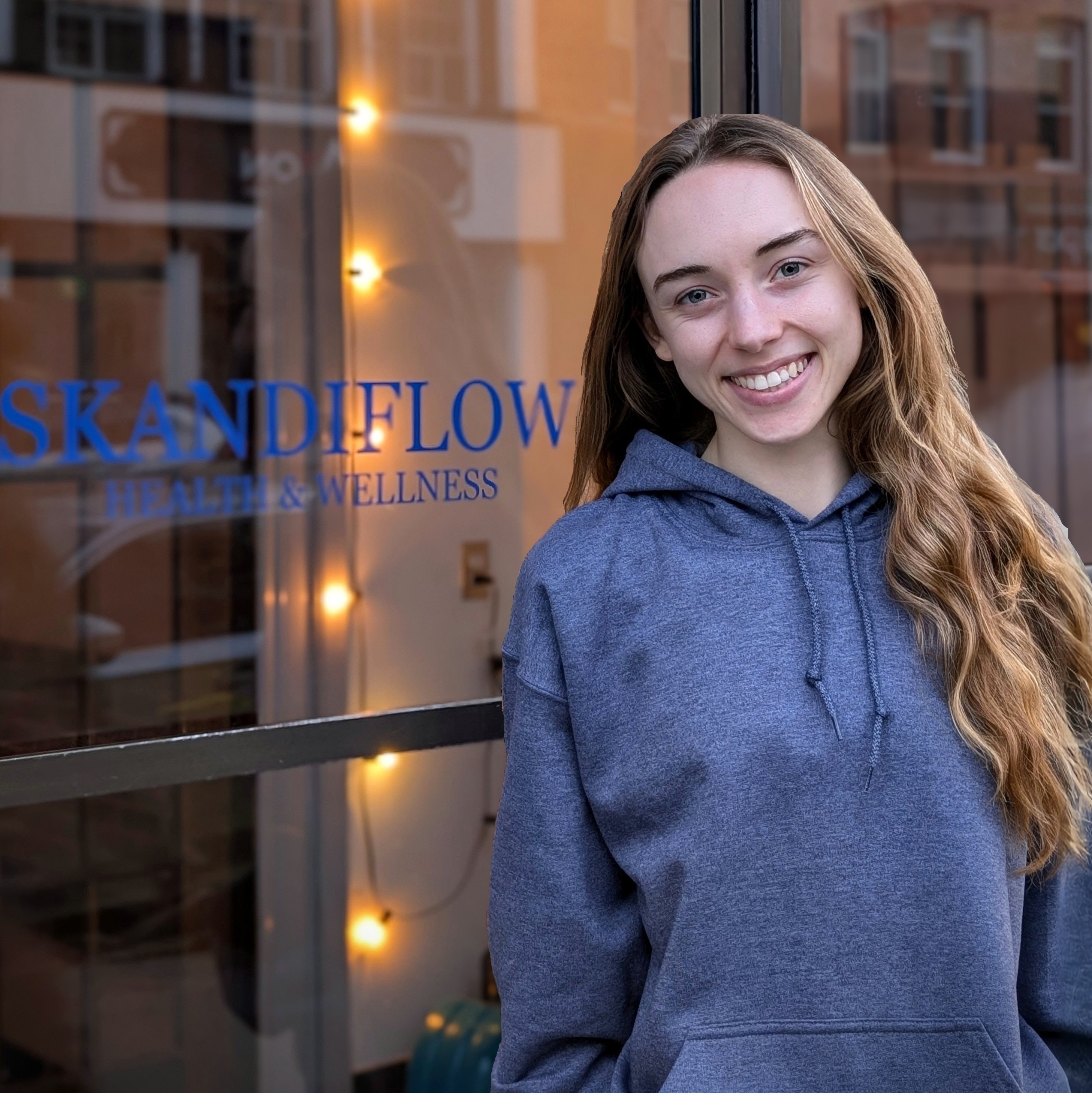 A young woman smiling in front of a business window displaying the name 'SKANDIFLOW HEALTH & WELLNESS' with decorative lights in the background.