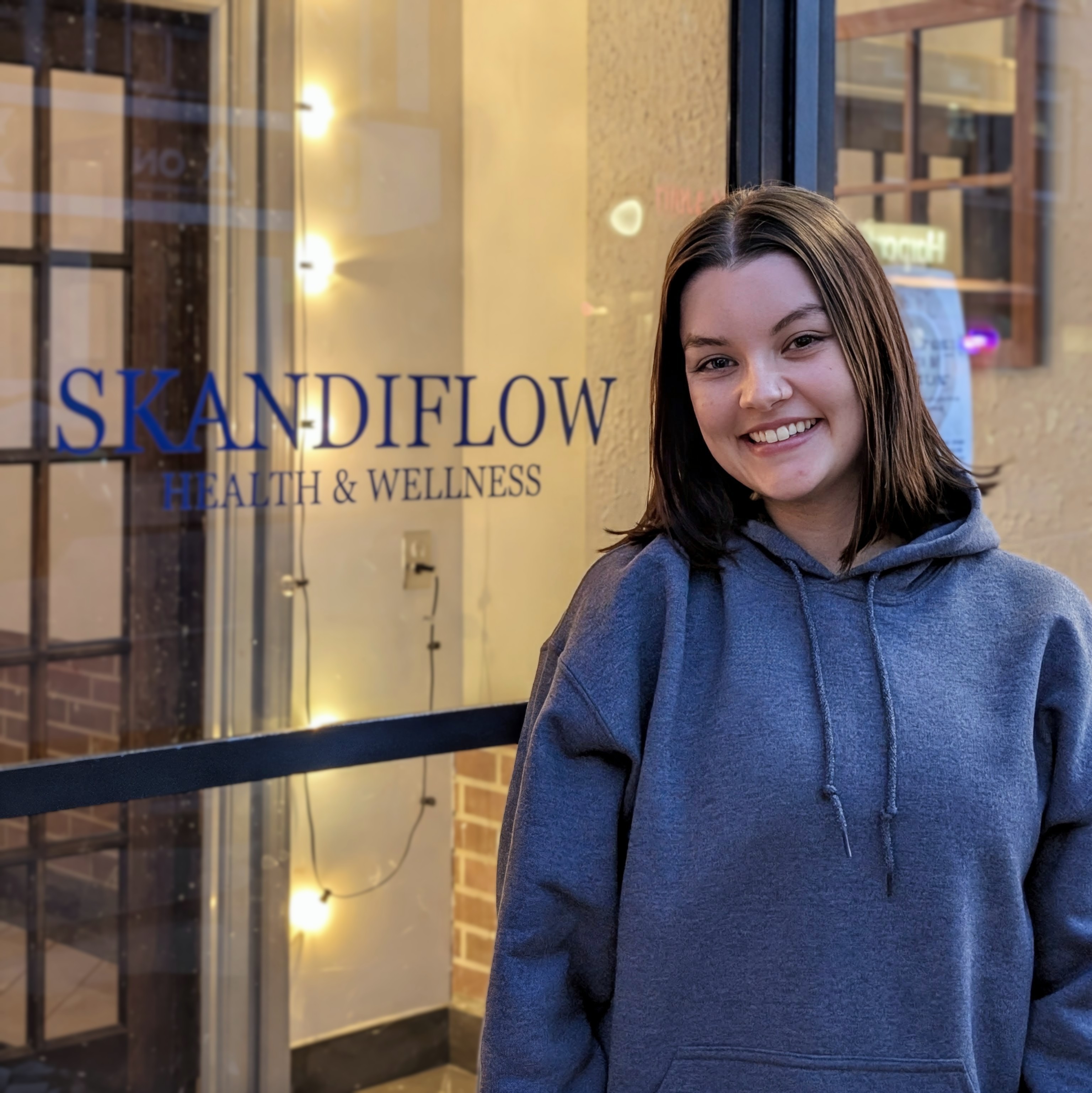 A smiling young woman stands outside a health and wellness store called 'SKANDIFLOW HEALTH & WELLNESS', wearing a gray hoodie.