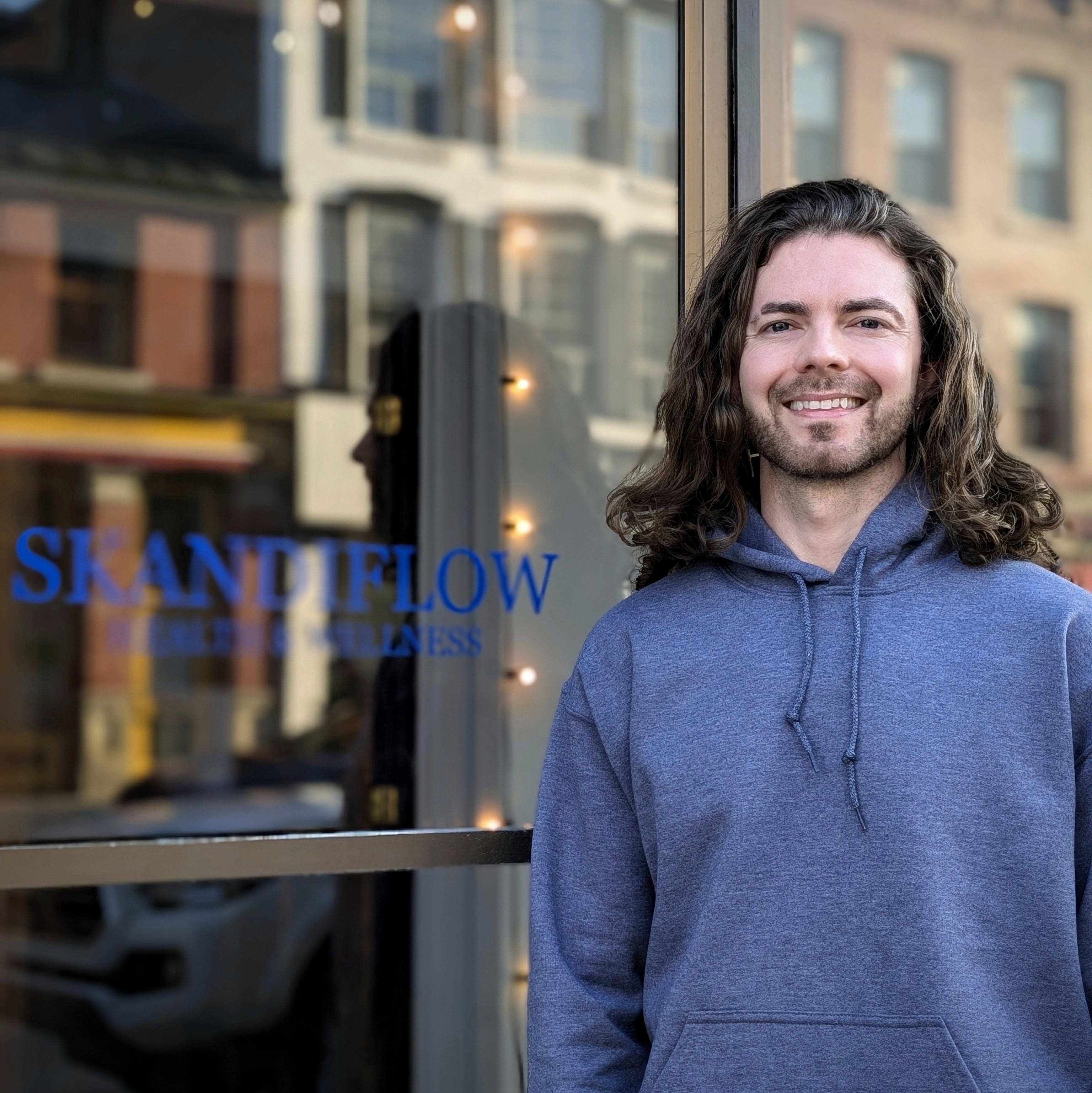 A smiling man with long hair stands outside a wellness store called 'SKANDIFLOW,' with large windows and a friendly atmosphere.