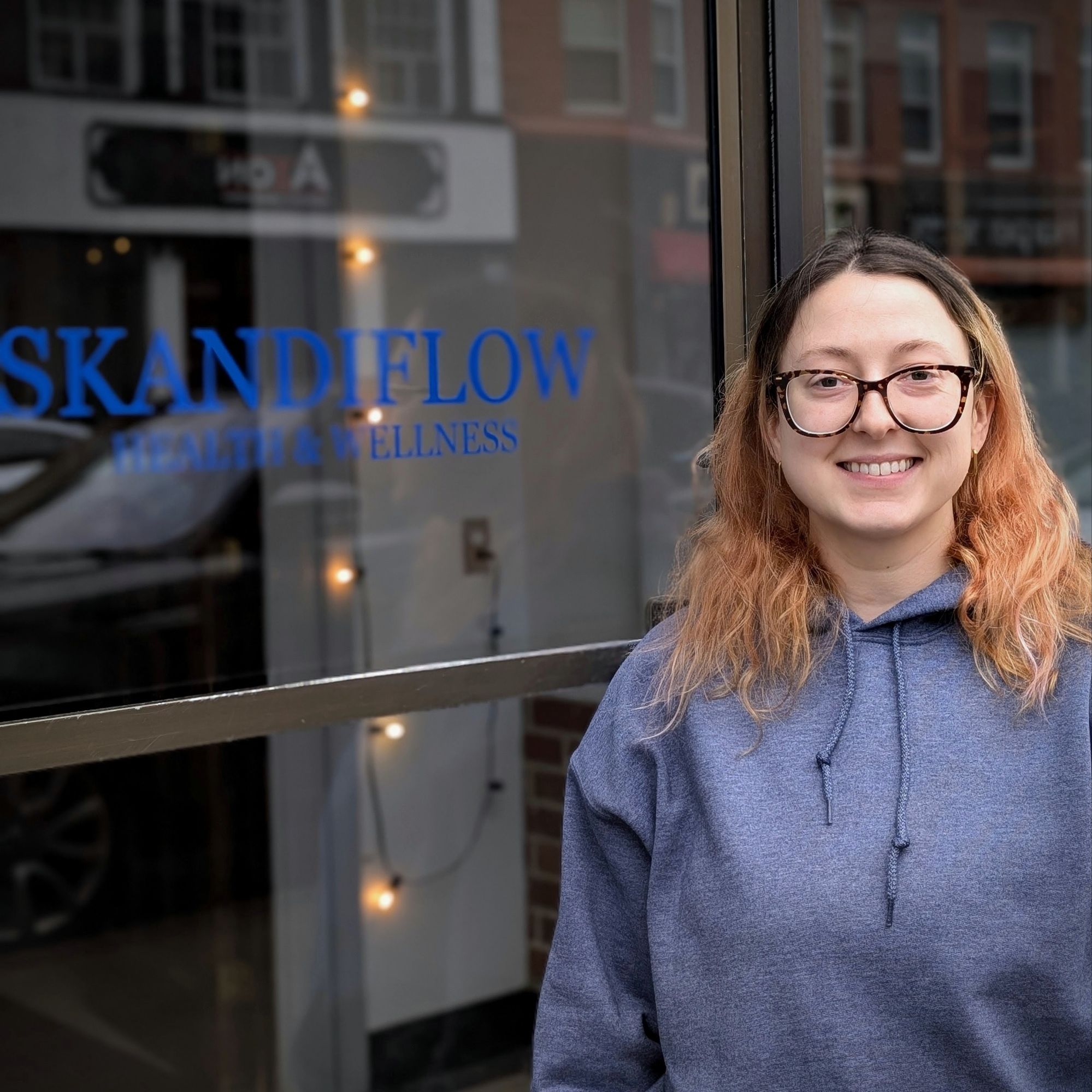 A smiling individual stands outside a business with a sign that reads 'Skandiflow Health & Wellness.' The person is wearing a gray hoodie and has long, wavy hair with lighter highlights.