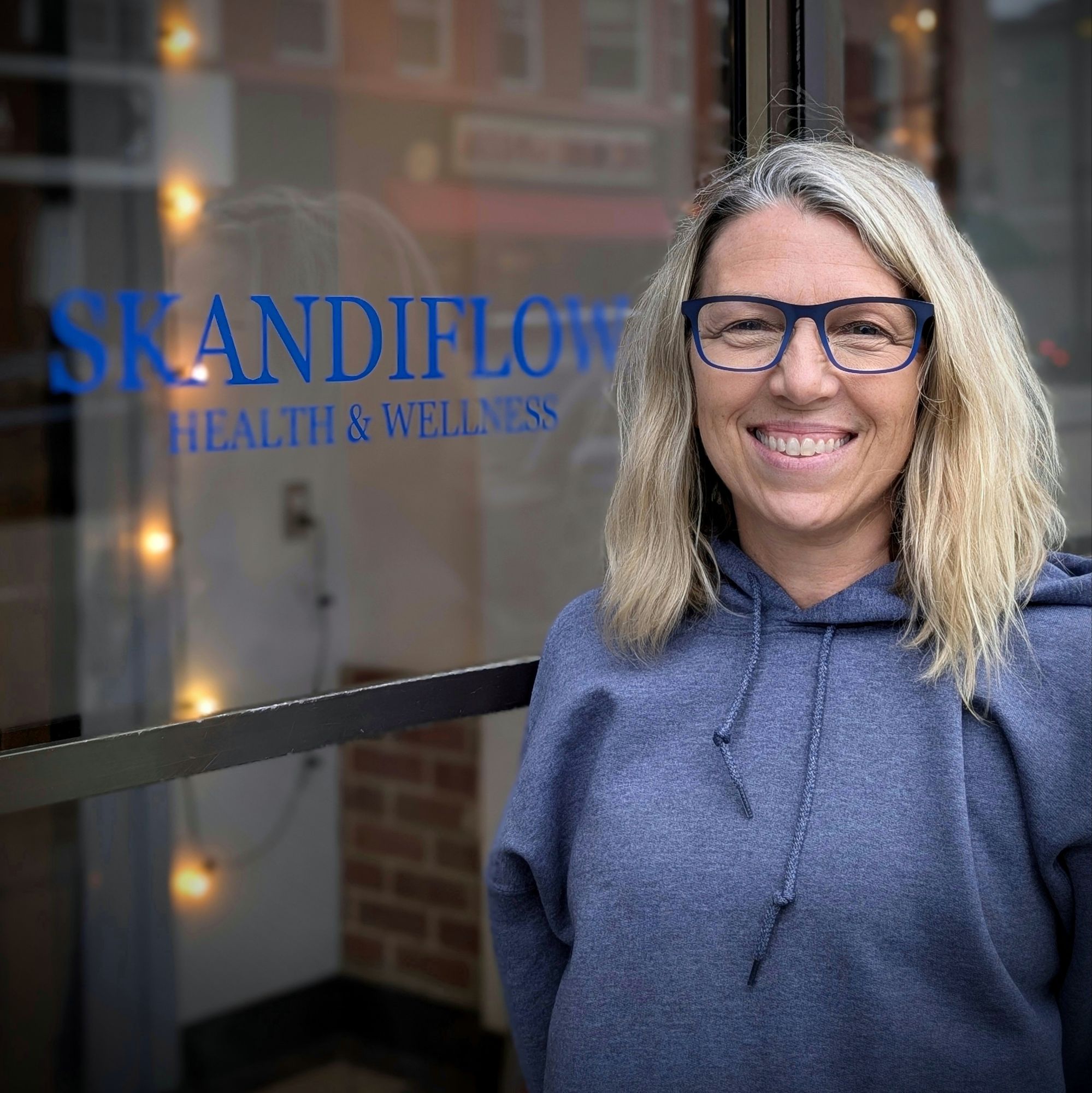 A smiling woman wearing a hoodie stands in front of a glass window displaying the sign 'SKANDIFLOW HEALTH & WELLNESS'.