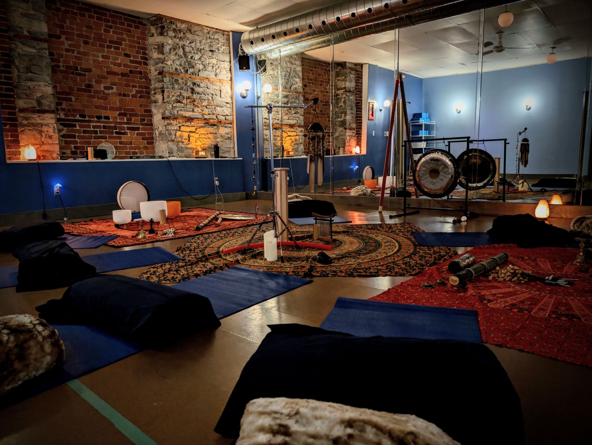 A serene meditation space featuring black cushions and colorful rugs, adorned with various musical instruments, including singing bowls and gongs, under warm lighting.